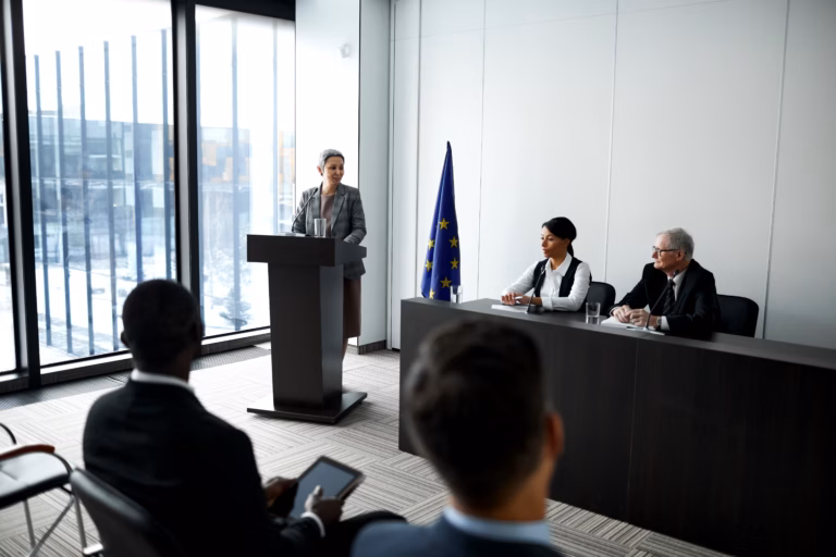 A woman stands at a podium, speaking to an audience in a well-lit conference room