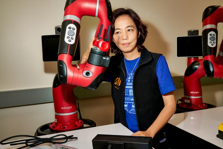 Fei-Fei Li stands beside a red robotic arm in a Stanford robotics lab.