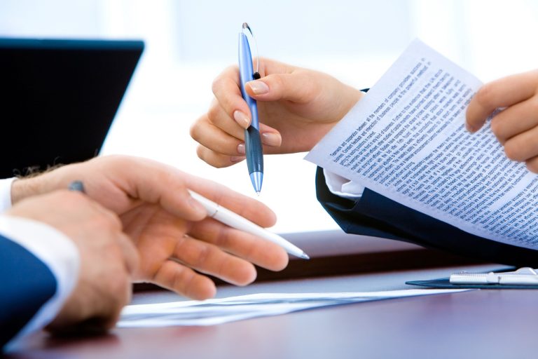 Hands holding pens and reviewing a printed document beside a laptop