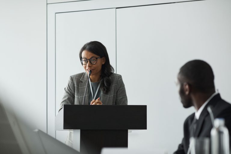 African American businesswoman delivering a speech at a conference podium with a colleague listening