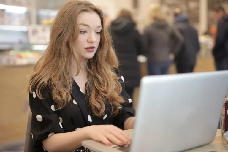 A focused woman in a polka dot shirt is typing on a laptop in a bustling cafe