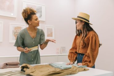 Two women discuss clothing samples at a worktable in a small fashion studio.